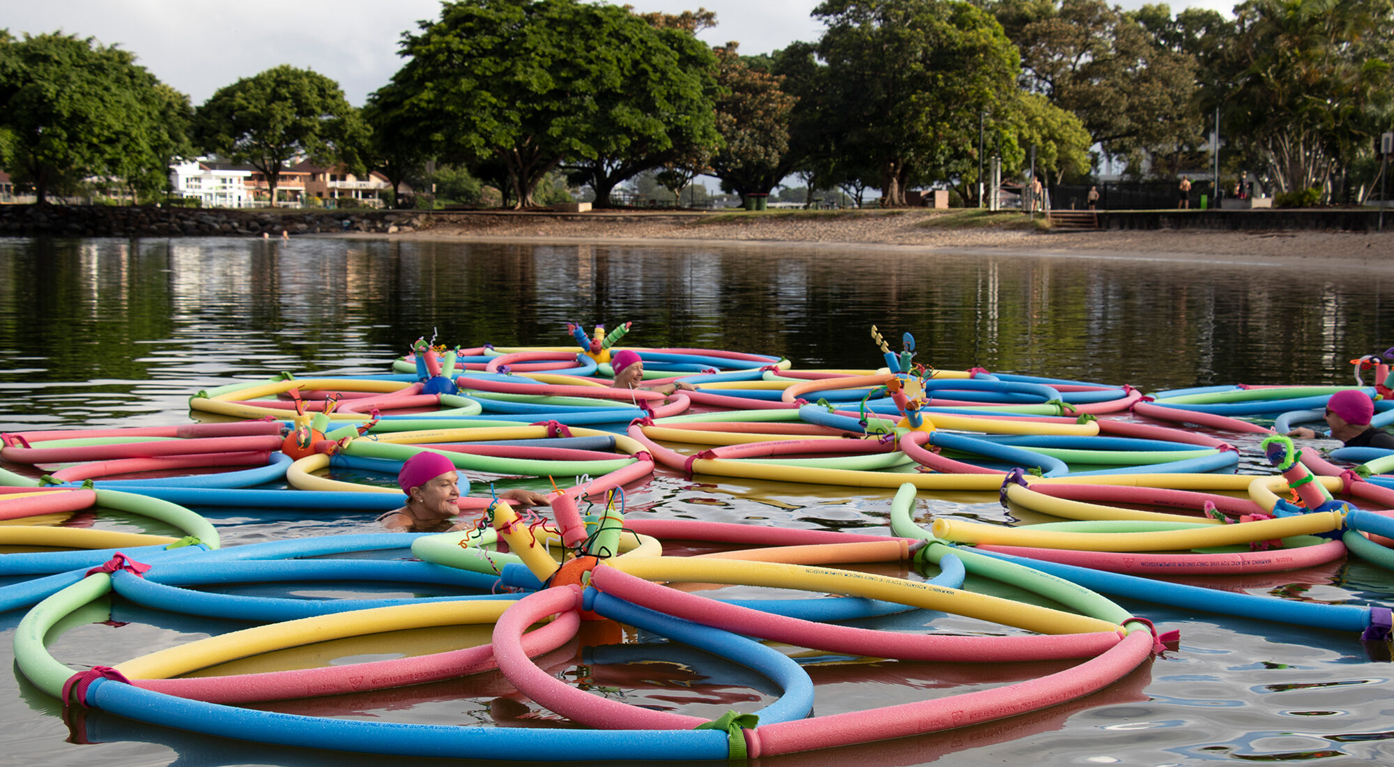 Pool toys floating in a river. There are people in swimming caps in the water, too.